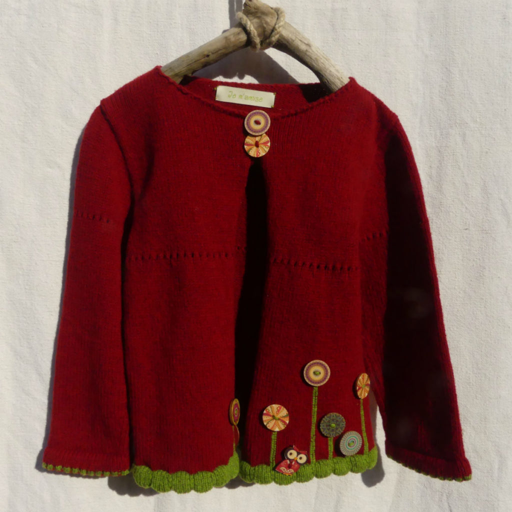 Cardigan manche longue de couleur rouge foncé avec une bordure pour les finitions de couleur verte . des boutons bois motif fleur au bas du cardigan.